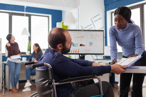 Black manager woman working with financial documents checking graphs talking with paralysed team leader with disabilities, reading raports sitting in wheelchair in start-up business office.