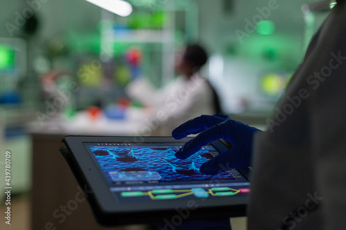 Scientist researcher doctor analyzing genetic mutation test using tablet while her collegue typing biological expertise on computer in background. Medical team working in microbiology laboratory.