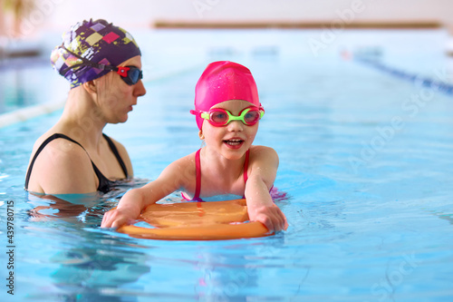 Portrait of mother or coach learning to swim with flutter board little girl at the public swimming-pool. Space for text
