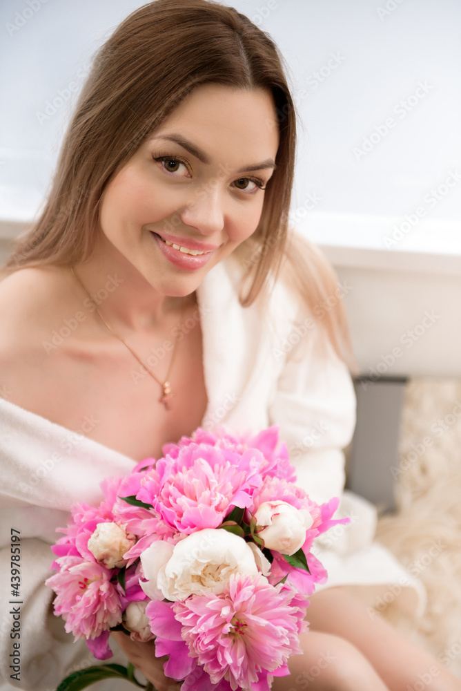 Beautiful girl in a white bathrobe with a bouquet of pink peonies near the bath.