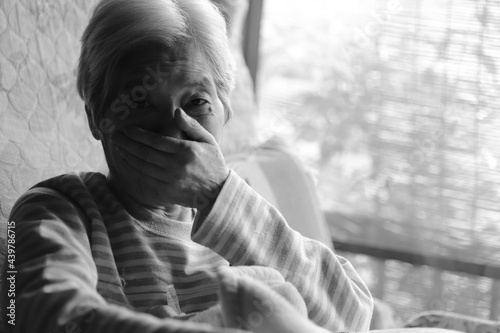 介護ベッドでくつろぎながら口元を手で覆う高齢女性 © michikodesign