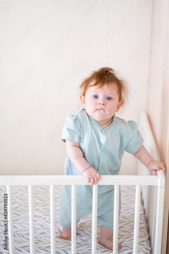 A toddler with red hair is holding onto the side of the crib. Danger for the child to fall out of bed. The baby learns to stand at the support. A toddler with red hair is holding onto the side of the crib. Danger for the child to fall out of bed. The baby learns to stand at the support.