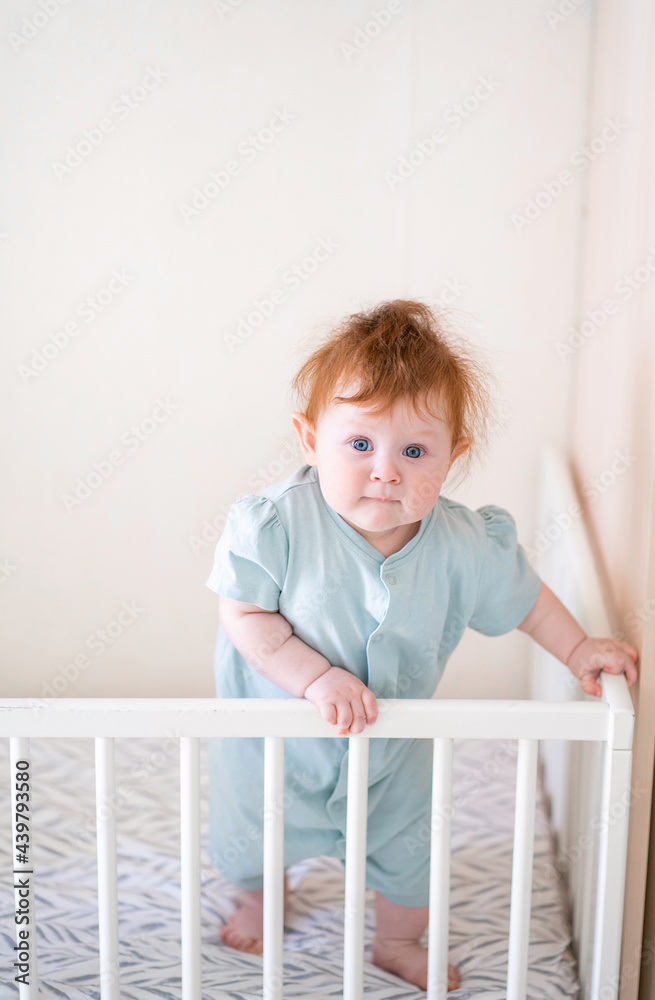 A toddler with red hair is holding onto the side of the crib. Danger for the child to fall out of bed. The baby learns to stand at the support. A toddler with red hair is holding onto the side of the crib. Danger for the child to fall out of bed. The baby learns to stand at the support.