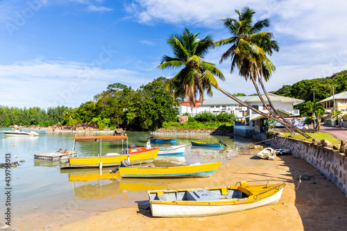Many colorful small wooden boats moored at Baie Sainte Anne bay on Praslin Island, with palm trees and tropical plants around, white sandy beach, and Sainte Anne village in the background.