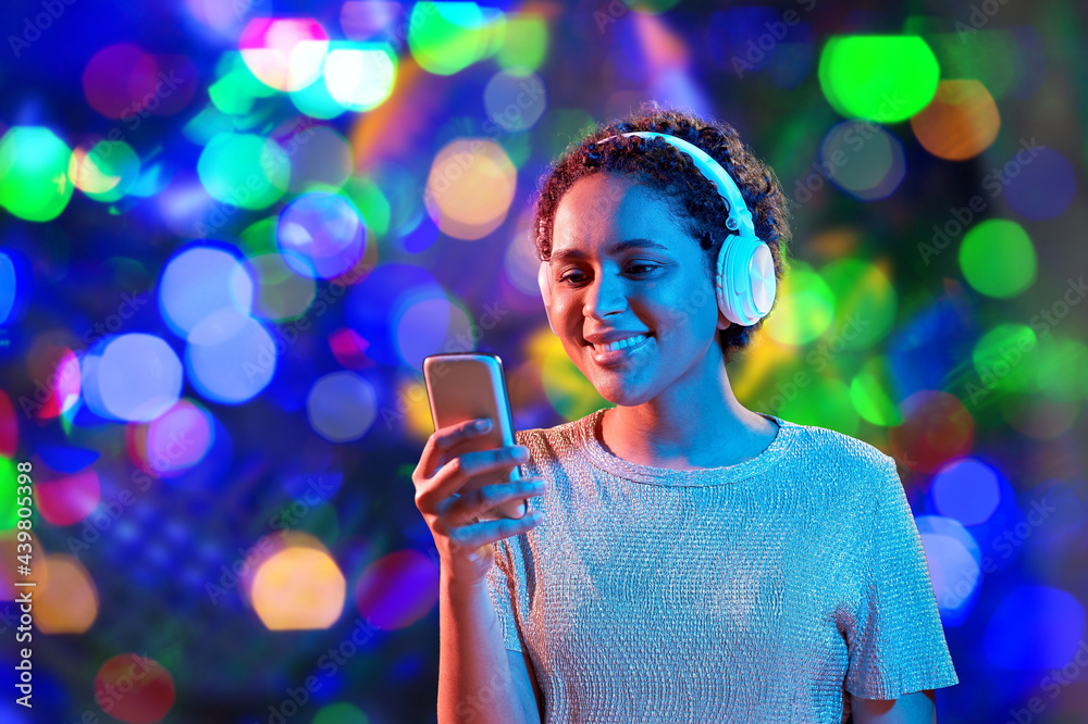 nightlife, fashion and people concept - happy young african american woman with smartphone and headphones in neon lights over bokeh background