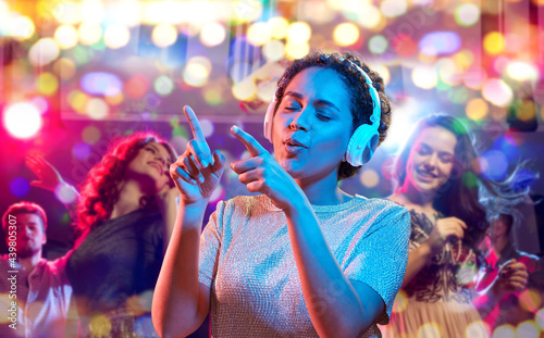 nightlife, technology and people concept - happy young african american woman in headphones listening to music and dancing in neon lights over nightclub background