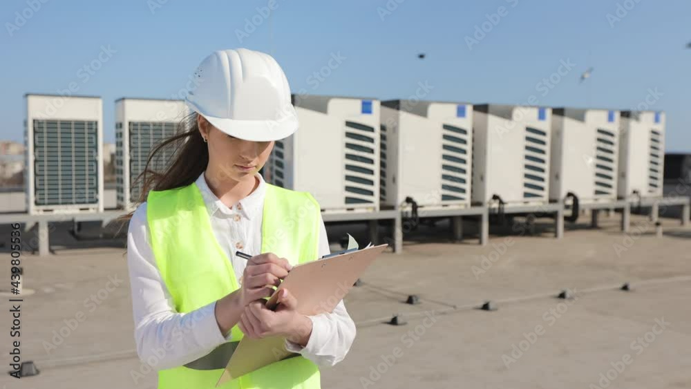 Happy woman engineer is making notes in documents and looking at the camera. She is smiling. Strong wind is blowing. She is wearing work clothes and a hard hat. Air conditioning systems in the