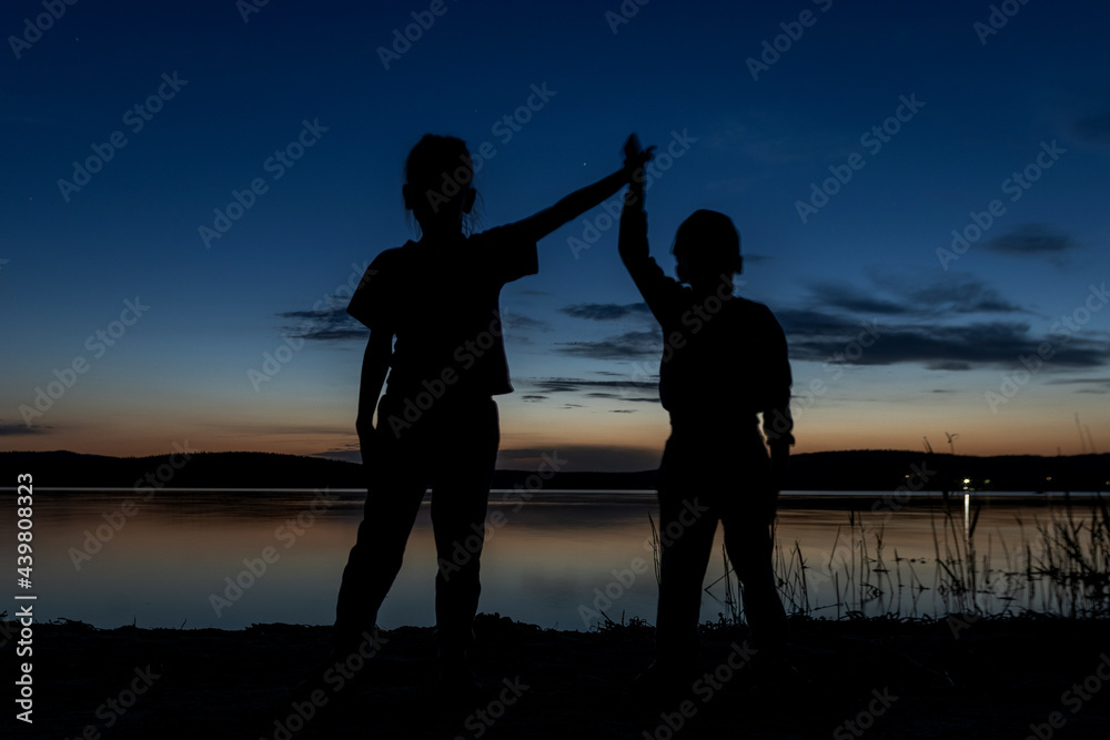 Silhouette of two children standing with their hands up on the shore of the lake at sunset. Family vacation by the lake, camping