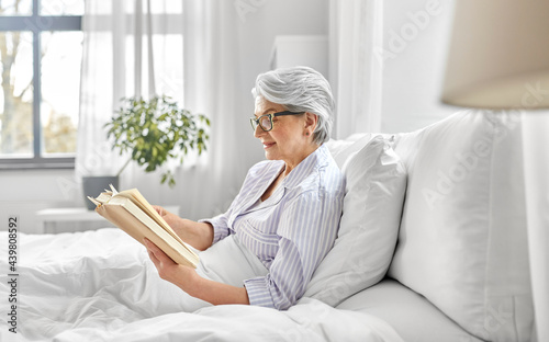 old age, leisure and people concept - happy smiling senior woman in glasses reading book in bed at home bedroom