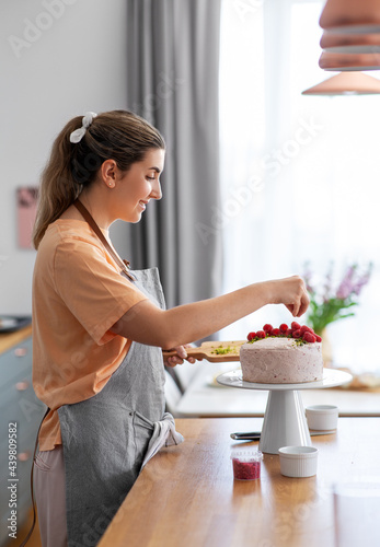 culinary, baking and cooking food concept - happy smiling young woman decorating cake with raspberries on kitchen at home