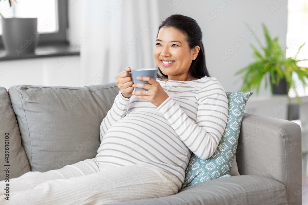 pregnancy, rest, people and expectation concept - happy smiling pregnant asian woman with mug sitting on sofa at home and drinking tea