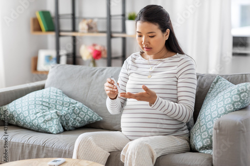 pregnancy, health and glycemia concept - pregnant asian woman checking blood sugar level with glucometer and lancing device at home
