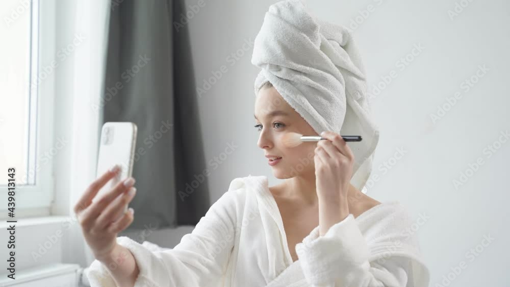 Caucasian woman using makeup brush and smiling in front of small mirror, at home, sitting on bed. Morning skin care beauty routine, self care. Caucasian woman using makeup brush and smiling in front of small mirror, at home, sitting on bed. Morning skin care beauty routine, self care.