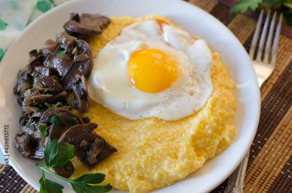 Corn porridge, fried egg, fried mushrooms with onions in a plate on a wicker bamboo napkin.