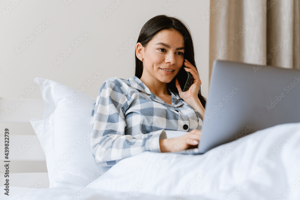 Young woman talking on cellphone and using laptop while lying in bed Young woman talking on cellphone and using laptop while lying in bed