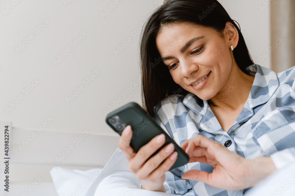 Young hispanic woman smiling and using cellphone while lying in bed Young hispanic woman smiling and using cellphone while lying in bed