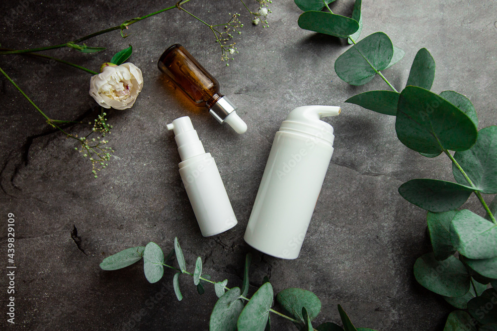 bottles with serum and jars of cosmetic creams on a gray background with eucalyptus sprigs and green leaves.