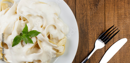 Tagliatelle pasta served with bechamel sauce and basil leaves. Pasta with white sauce. Served on white plate. Top view photo. Fork and knife on the right.