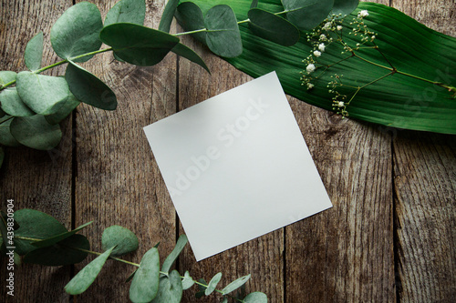 blank white sheet with place for text on a brown wooden background with plant leaves, flowers and eucalyptus sprigs.
