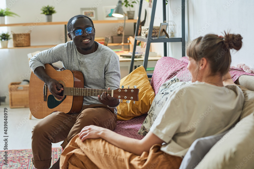 African young man playing guitar and singing songs for his girlfriend on the sofa in the room