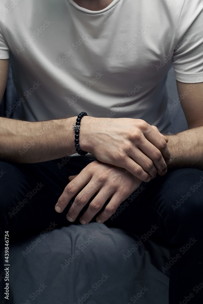 Cropped shot of a man with beaded bracelet made of black ornamental stone and decorated with little crown with crystals. Man in white t-shirt and dark jeans is sitting with crossed hands. 