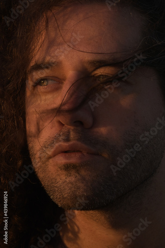 Handsome light-eyed man with curlers and hair on his face looking to the side. Isolated portrait with a soft warm light on the face of a beautiful sunset