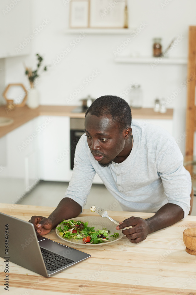 African man working on laptop online and eating fresh salad in the kitchen