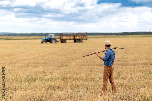 Canvas-taulu A farmer holds a pitchfork and looks at a blue tractor in a field