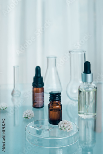 Brown bottle on glass cup with white flower and drop of liquid closeup with blurred flasks and bottles in the background, all on glassy surface. Natural essences and extractions concept.