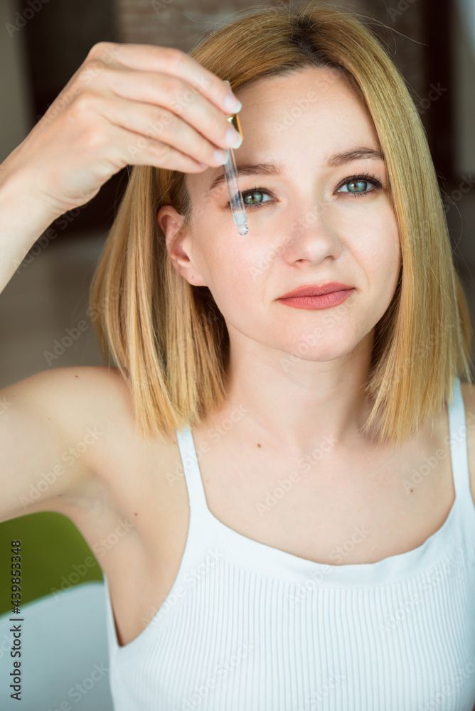 Fototapeta premium A beautiful young woman holds a glass-dropper with cosmetical serum upon her face. Skin treatment and cosmetology concept.