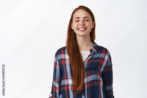 People and lifestyle. Happy beautiful redhead girl with long natural hair, freckles and no make up, dreaming of something, imaging and smiling with closed eyes, white background © Cookie Studio