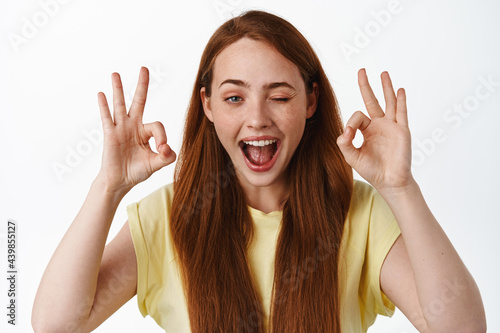 Close up portrait of redhead positive girl, winking and showing okay OK sign, no problem, praise good thing, well done, great job, standing over white background