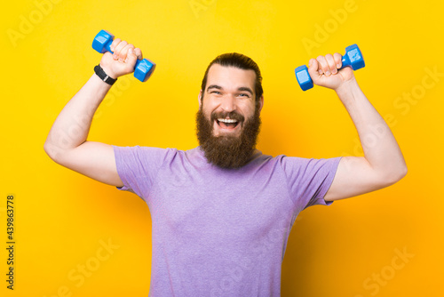 Obraz na plátně Portrait of happy cheerful bearded man working with small dumbbells over yellow