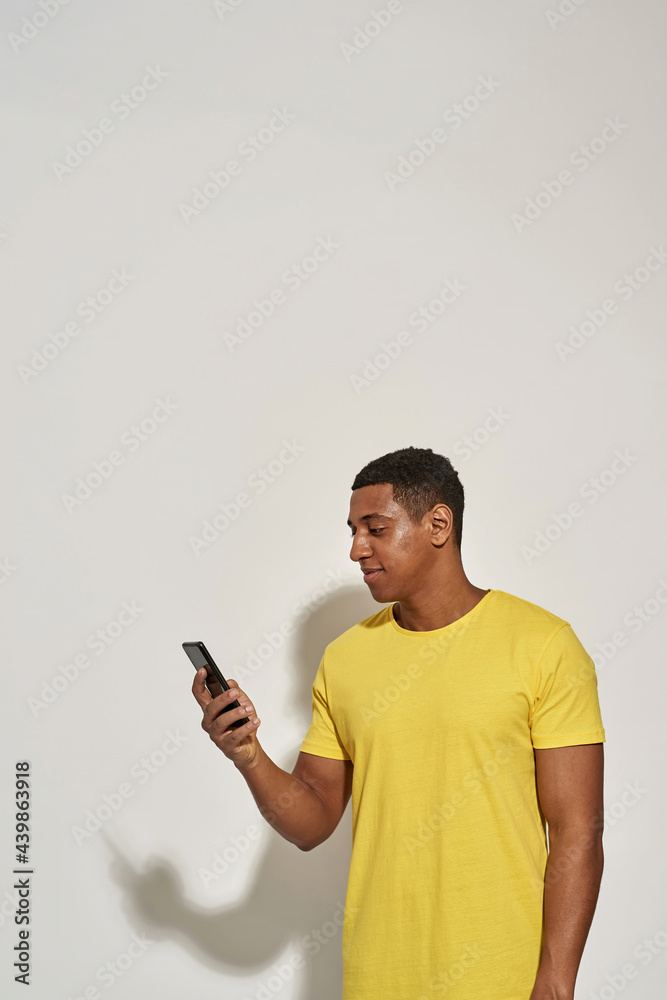 Focused young man in casual yellow t shirt looking busy, holding and using his smartphone while standing isolated over gray background Focused young man in casual yellow t shirt looking busy, holding and using his smartphone while standing isolated over gray background