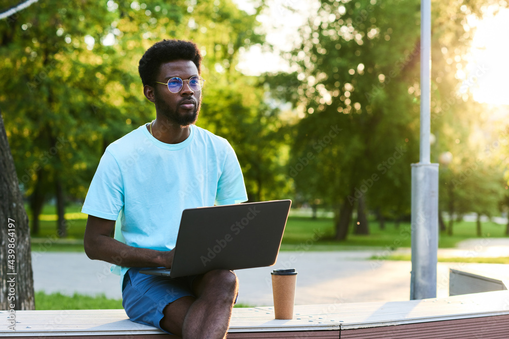 Serious African businessman working online on laptop while sitting in the park outdoors Serious African businessman working online on laptop while sitting in the park outdoors