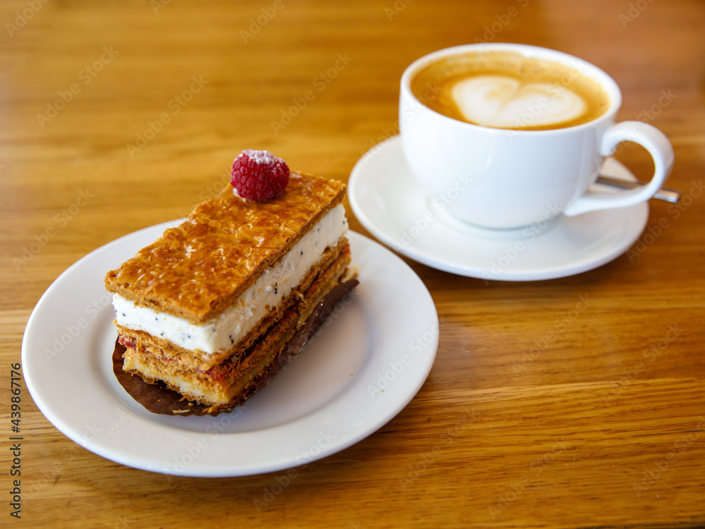 Berry milfey cake on a white plate with cup of cappuccino coffee on wooden table, horizontal, side view. Breakfast or snack or launch in cafe Berry milfey cake on a white plate with cup of cappuccino coffee on wooden table, horizontal, side view. Breakfast or snack or launch in cafe