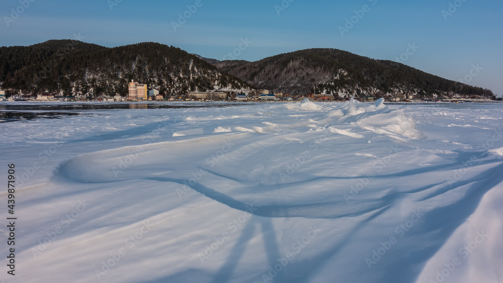 The frozen lake is covered with snow. Shadows on the relief of snowdrifts. Mountain range against the blue sky. Village on the coast. Baikal The frozen lake is covered with snow. Shadows on the relief of snowdrifts. Mountain range against the blue sky. Village on the coast. Baikal