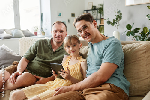 Portrait of happy gay family smiling at camera while sitting on sofa with child and using tablet pc