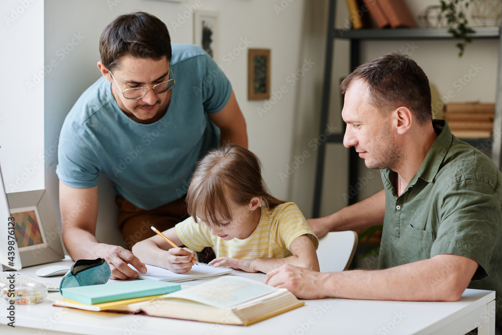 Little girl sitting at the table and doing her homework while gay parents helping her Little girl sitting at the table and doing her homework while gay parents helping her