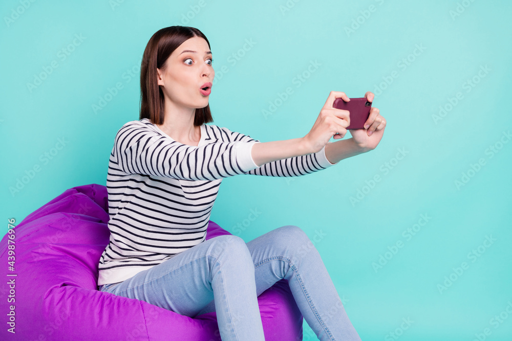 Photo of shocked addicted lady sit bean bag play video game phone wear striped shirt isolated on teal color background Photo of shocked addicted lady sit bean bag play video game phone wear striped shirt isolated on teal color background