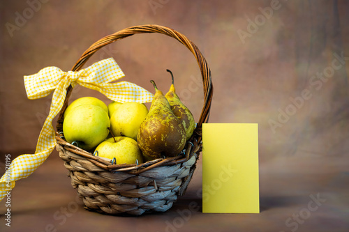 Cesto con frutta pieno di pere e mele con un fiocco giallo e un biglietto di auguri giallo accanto. Alimentazione, invito, ringraziamento, salute. photo