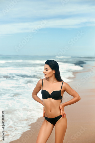 Portrait of young woman in black bikini on tropical beach looking at camera. Beautiful latin girl in swimwear with copy space. Sexy tanned body. © verona_studio