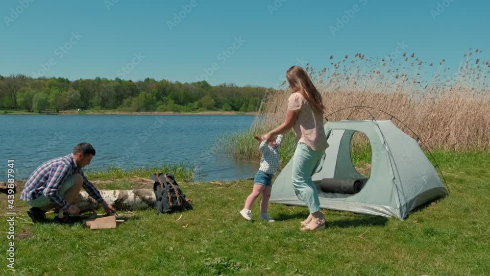 Family is resting on the bank of the river with a tent. Father mother and little daughter collect firewood Family is resting on the bank of the river with a tent. Father mother and little daughter collect firewood