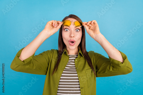 Photo of shocked astonished young woman raise sunglass reactionunexpectedd news isolated on blue color background