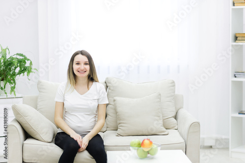 Cropped photo of pregnant woman sitting on white sofa. © Andrii