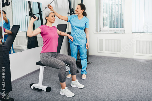 Mature woman sitting on sports bench while using pull rope exercise machine for rehabilitation near her rehabilitologist at medical sport center photo