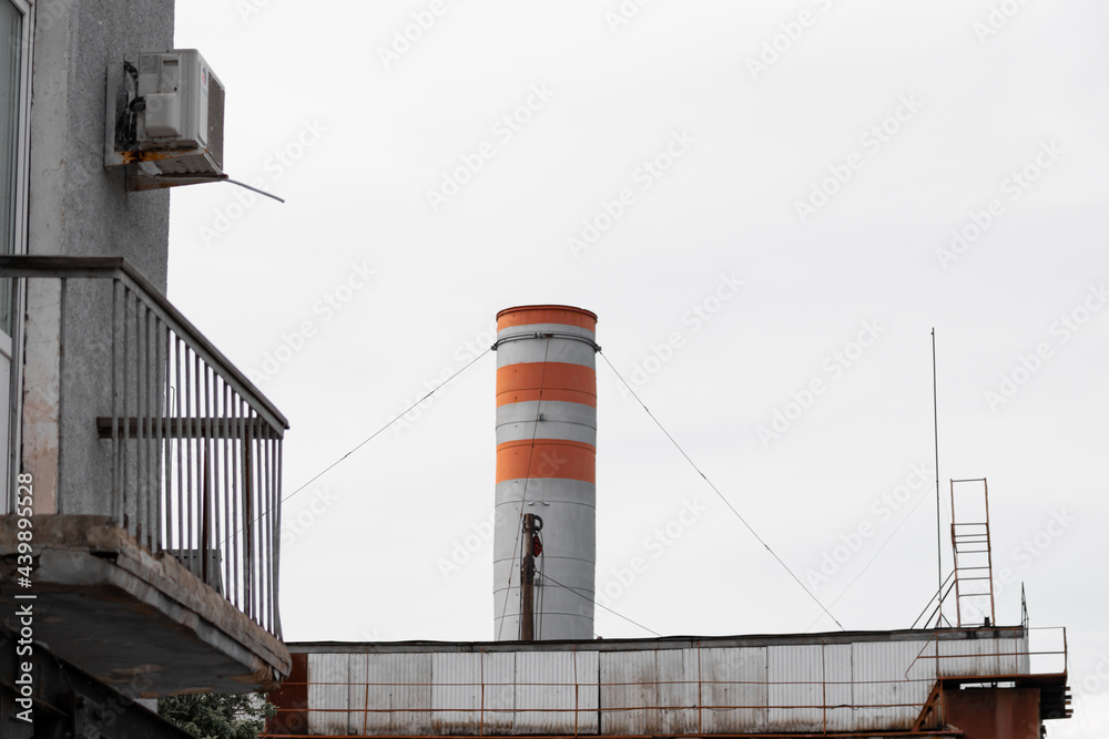 A large high industrial chimney rises above the factory building against the background of a clear white sky in the factory district of the city in the industrial zone. Copy space. High quality photo A large high industrial chimney rises above the factory building against the background of a clear white sky in the factory district of the city in the industrial zone. Copy space. High quality photo