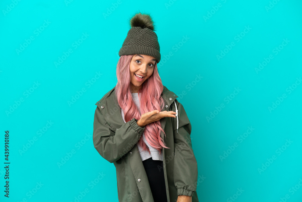 Young woman with pink hair wearing a rainproof coat isolated on blue background presenting an idea while looking smiling towards Young woman with pink hair wearing a rainproof coat isolated on blue background presenting an idea while looking smiling towards