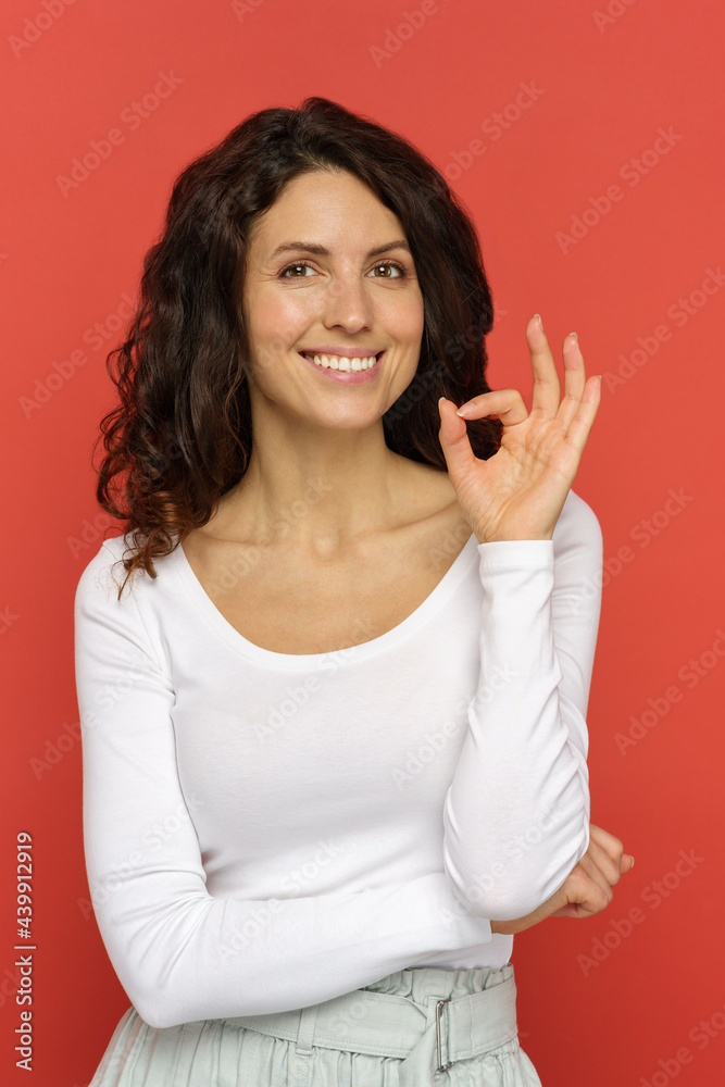Portrait of happy cheerful smiling female showing okay gesture. Young curly woman in white outfit hold ok finger sign over red studio wall background. Success, approval and gesturing language concept Portrait of happy cheerful smiling female showing okay gesture. Young curly woman in white outfit hold ok finger sign over red studio wall background. Success, approval and gesturing language concept