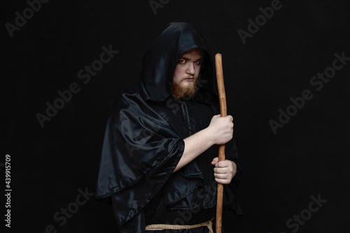 A monk in a black hood with a staff on a dark background, a gloomy expression, a medium plan, witchcraft and curses.